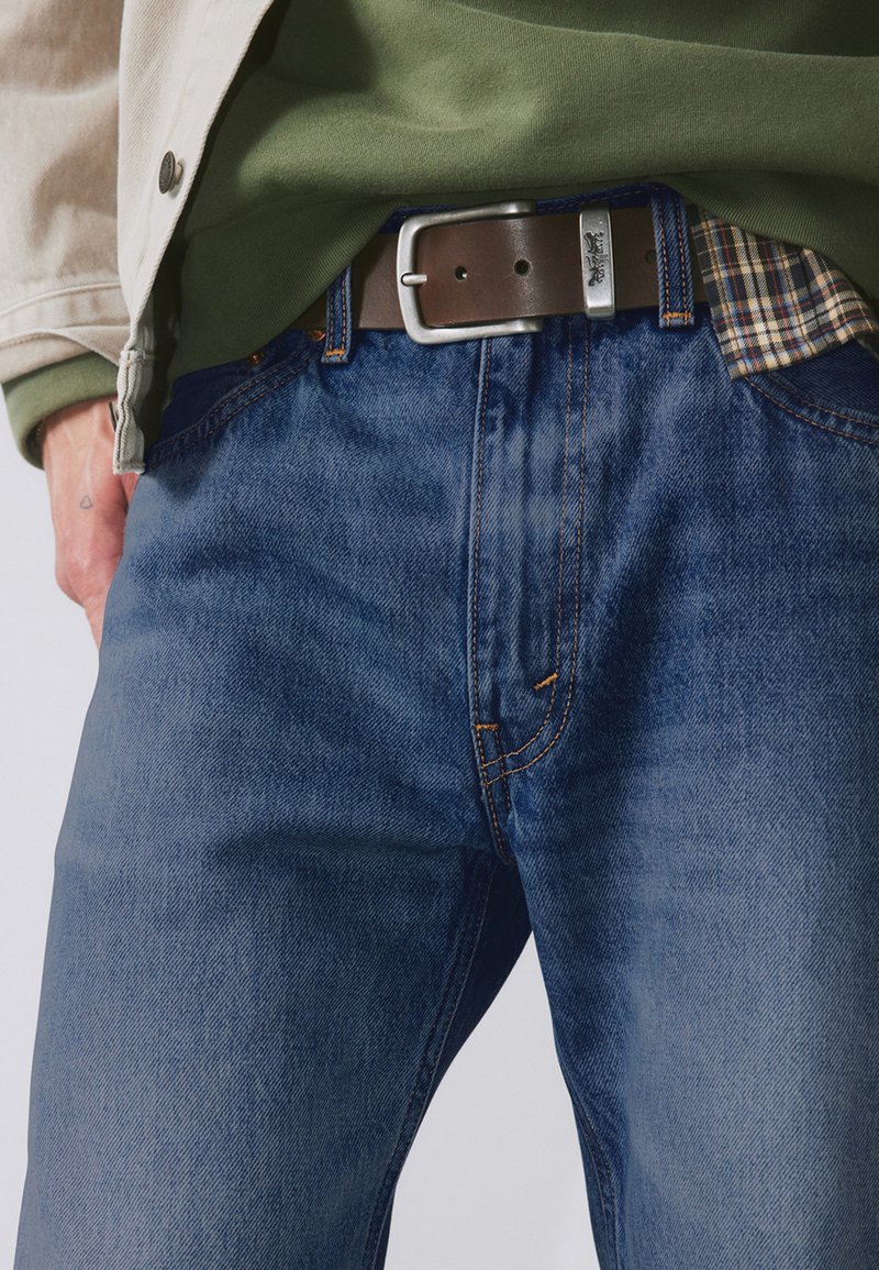 Jean en denim bleu foncé avec surpiqûres orange, assorti à une ceinture en cuir marron avec une boucle argentée et une chemise à carreaux en dessous.
