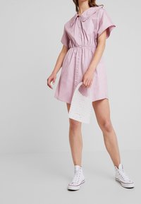 Abito rosa a maniche corte con colletto, vita cin cin e chiusura a bottoni, abbinato a una borsa in crochet bianca e sneakers bianche alte.