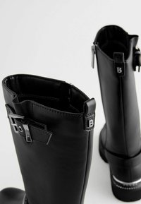 Schwarze Lederstiefel bis zum Knie mit glatter Textur, versehen mit einem silbernen Schnallenakzent und einem Reißverschluss, der ein elegantes Design unterstreicht.