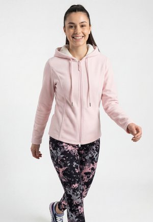 Jonge vrouw die glimlacht en loopt, draagt een lichtroze zip-up hoodie, zwart-roze patterned legging en sportschoenen.
