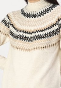 Strickpullover in Creme mit einem strukturierten Einsatzdesign, das beige und schwarze Zickzackmuster aufweist. Hoher Kragen und lockere Passform.