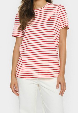 Mujer vistiendo una camiseta blanca de manga corta con rayas rojas horizontales y pequeños detalles de corazones rojos, combinada con pantalones blancos.