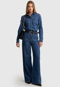 Monos de mezclilla en azul oscuro, con mangas largas, dos bolsillos en el pecho, una cintura ceñida y pantalones de pernera ancha. Bolso negro con cadena.