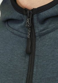 Mørkeblå zip-up hoodie lavet af blødt stof. Har en sort lynlås med et tekstureret trækstykke og en justerbar snor i hætten.