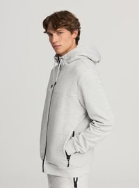 Felpa grigia con zip frontale realizzata in tessuto morbido, con cappuccio, dettagli della zip neri e tasche laterali con zip. Silhouette casual, aspetto strutturato.