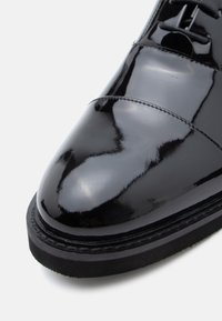 Svart lakksko med rund tå, flat såle og detaljert søm. Høyglanset finish fremhever dens glatte tekstur.