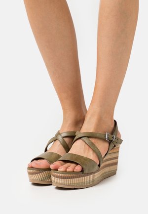 Sandalias de cuña verde oliva con tiras cruzadas, puntera abierta y textura trenzada sobre una base de plataforma. Incluye detalles de hebilla metálica.