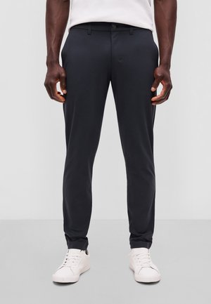 Mand iført sorte slim-fit bukser og hvide sneakers, stående mod en ensfarvet lys baggrund.