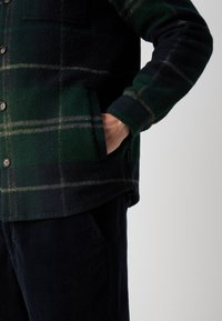 Roheline ja tume ruuduline flanellshirt koos nööpidega, millel on rindade tasku ja pehme tekstuur. Sobib siniste vähipükstega.