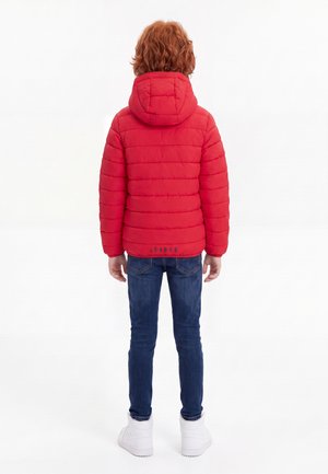 Enfant aux cheveux roux bouclés portant une doudoune rouge à capuche, un jean bleu et des baskets blanches, debout de dos sur un fond blanc.