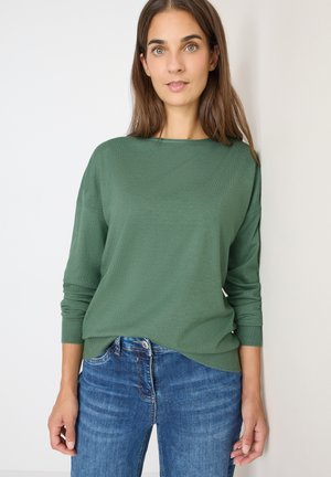Maglione verde a maniche lunghe con vestibilità comoda, lavorazione a maglia testurizzata e polsini a costine, abbinato a jeans blu, su uno sfondo neutro.