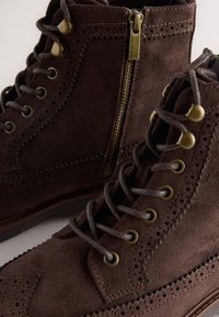 Hnedé semišové členkové topánky s brogue zdobením, bočným zipsom a bronzovými doplnkami. Dizajn dopĺňajú šnúrky a dekoratívne perforácie.