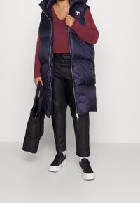 Marineblaue Steppweste mit Kapuze und goldenen Reißverschluss, kombiniert mit einem burgunderfarbenen Pullover, schwarzen Kunstlederhosen und schwarzen Sneakers.