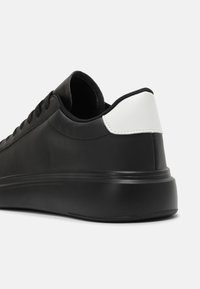 Černá nízká sneakers s tlustou podrážkou, černými tkaničkami a bílým zpevněním na patě, zobrazená ze zadního úhlu na bílém pozadí.
