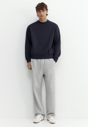Junger Mann steht an einer weißen Wand und trägt einen dunkelblauen Pullover, hellgraue Jogginghosen und weiße Sneakers mit hellbraunen Sohlen.