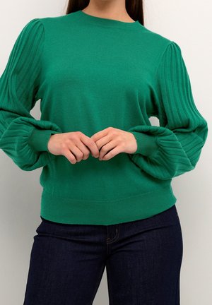 Mujer con un suéter verde de mangas abullonadas plisadas, combinado con jeans azul oscuro, con las manos entrelazadas al frente.