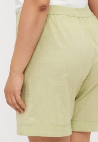 Pantalones cortos verde claro fabricados con una tela texturizada, con una cinturilla elástica y dobladillos enrollados, presentando un ajuste relajado visible desde el lado.