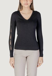 Maglione nero a V con maniche lunghe, caratterizzato da pannelli in pizzo sui lati. Materiale morbido e design aderente. Abbinato a pantaloni chiari.