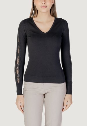 Maglione nero a V con maniche lunghe, caratterizzato da pannelli in pizzo sui lati. Materiale morbido e design aderente. Abbinato a pantaloni chiari.