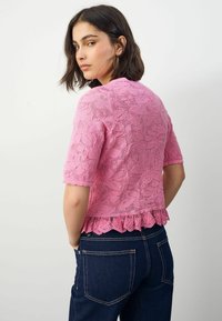 Růžový krajkový top s krátkými rukávy, květinový vzor, vroubkovaný lem, vrstvená textura, kombinovaný s tmavými džíny. Detailní pohled zezadu.