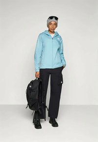 Svetlomodrá bunda na zips, čierne cargo nohavice, čierny batoh a rukavice. Model s dôverou stojí s okuliarmi na očiach a predvádza vonkajšie oblečenie.