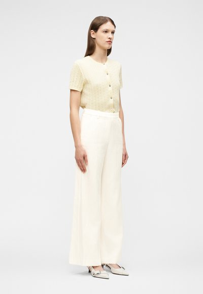 Γυναίκα όρθια, φοράει ένα ανοιχτό κίτρινο πουλόβερ με κοντά μανίκια και κουμπιά, λευκό παντελόνι με ψηλή μέση και φαρδιά μπατζάκια, και λευκά μυτερά παπούτσια με φιόγκους.