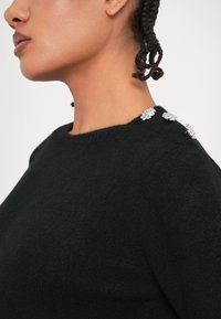 Sweater preto com decote redondo, apresentando três detalhes decorativos em prata no ombro, feito de um material suave e texturizado.