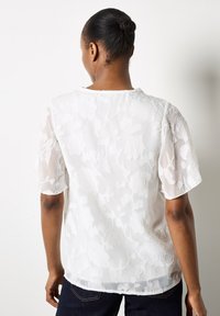 Weiße transparente Bluse mit floralem Muster, kurzen Ärmeln und einem runden Ausschnitt. Leichter Stoff fällt locker für eine entspannte Passform.