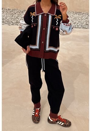 Femme portant un cardigan à motifs, un pantalon de jogging noir et des baskets à imprimé léopard avec des lacets rouges, tenant une pochette noire.