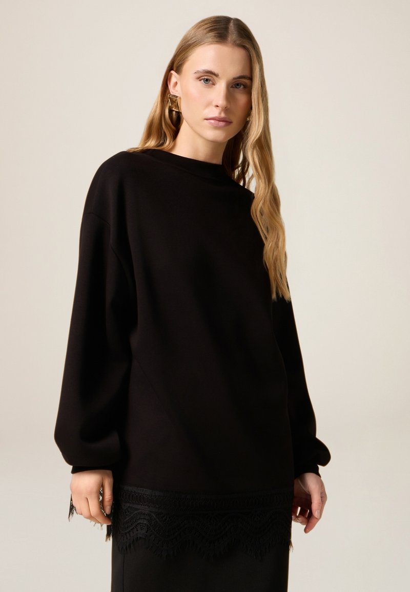 Sudadera negra de gran tamaño con mangas largas, que presenta un detalle de borde de encaje texturizado. Tela suave con un diseño relajado, ideal para superponer.