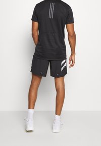 Camisa atlética negra con tres rayas verticales en la parte superior de la espalda. Combinada con pantalones cortos negros que presentan un patrón gráfico blanco. Zapatillas blancas.