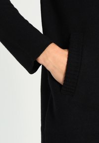 Musta neulottu vaatekappale, jossa on sivutasku, resorireunus ja sileä pinta. Käsi osittain näkyvissä taskussa.