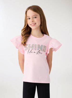 Uśmiechnięta dziewczyna z długimi włosami, ubrana w różową koszulkę z marszczonymi rękawami i napisem „SHINE like a star”, stoi na jednolitym białym tle.