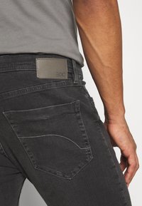 Vaqueros de mezclilla negra con un acabado sutilmente texturizado. Presentan un parche con el logotipo "EDC" y costuras curvas en los bolsillos traseros.