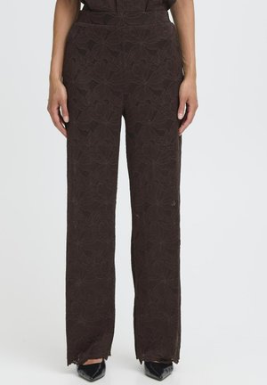 Pantalons larges à motif floral marron avec une finition texturée et une taille plate, présentant des ourlets effilochés et les mains du modèle sur les côtés.
