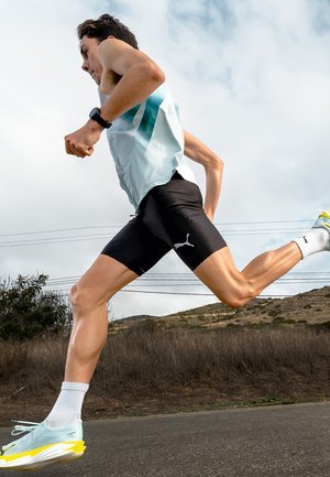 Hardloper in sportkleding en een smartwatch, vastgelegd halverwege een pas op een landweg met heuvels en elektriciteitsmasten op de achtergrond.