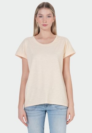 Jonge vrouw met steil blond haar, gekleed in een licht beige T-shirt met korte mouwen en lichtblauwe spijkerbroek tegen een effen achtergrond.
