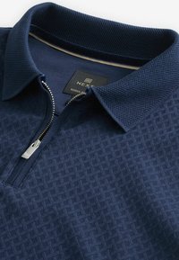 Tricou polo albastru marin cu un model texturat, guler din plasă și un fermoar pe jumătate în față. Materialul pare moale și respirabil, având un design casual.