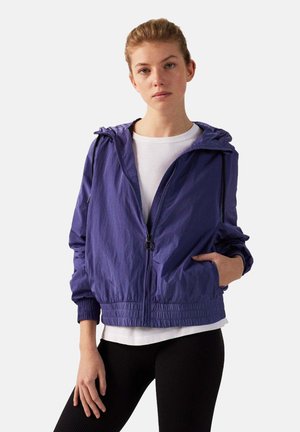 Paarse lichtgewicht jas met capuchon, rits aan de voorkant, elastische manchetten en een geraffineerde zoom; gedragen over een witte top en zwarte leggings.