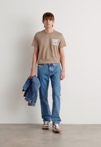 T-shirt beige à manches courtes avec une poche poitrine, associé à un jean bleu clair. Le modèle tient une veste en jean et porte des chaussures blanches et marron.