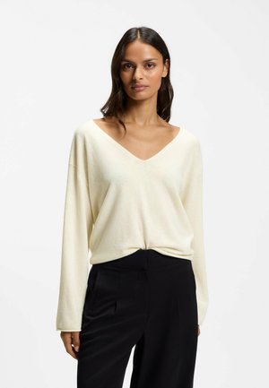 Femme aux cheveux mi-longs foncés portant un pull blanc ample à col en V et un pantalon noir taille haute sur un fond blanc uni.