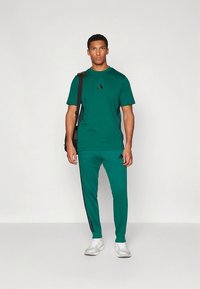 Πράσινο αθλητικό σύνολο που περιλαμβάνει μπλουζάκι με κοντό μανίκι και παντελόνι, και τα δύο με μαύρες λεπτομέρειες και λογότυπα. Ολοκληρώνεται με λευκά αθλητικά παπούτσια.