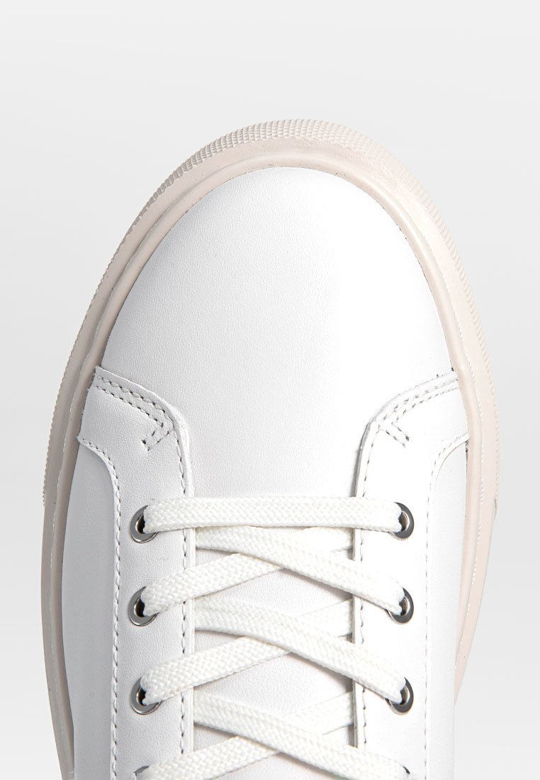 Zapatilla de cuero blanca con una puntera redonda y texturizada, cordones planos y una suela de goma de color claro. Presenta costuras decorativas y ojales de metal.