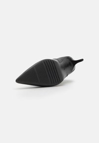 Sapata de salto alto preta com um design de bico fino. Material superior liso e sola de borracha texturizada com ranhuras horizontais para aderência.