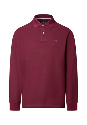 Hombre con una camisa polo de manga larga color burdeos, pantalones verdes a cuadros y zapatos de cuero marrón oscuro, de pie con las manos en los bolsillos.