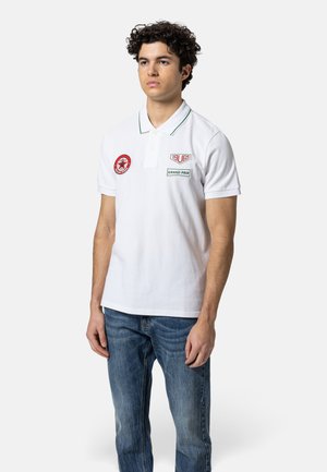 Junger Mann, der ein weißes Poloshirt mit Rennabzeichen und blaue Jeans trägt, steht vor einem schlichten hellen Hintergrund.