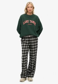 Zielona bluza z napisem "LAKE TAHOE", zestawiona z szerokimi spodniami w czarno-białą kratę. Modelka ma na sobie beżowe sneakersy.