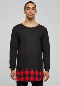 T-shirt noir à manches longues avec un col usé, superposé sur une chemise en flanelle à carreaux rouges et noirs, avec un ourlet arrondi.