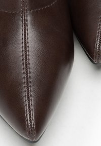 Detailní záběr tmavě hnědých kožených špičatých bot ukazujících detailní šití podél středního švu na světlém pozadí.