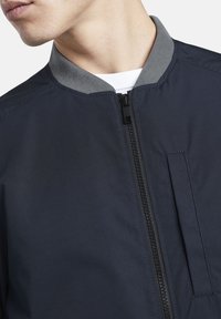 Námořnická bombardovací bunda s šedým límcem, doplněná předním zipem a boční kapsou. Hladká textura a minimalistický design.